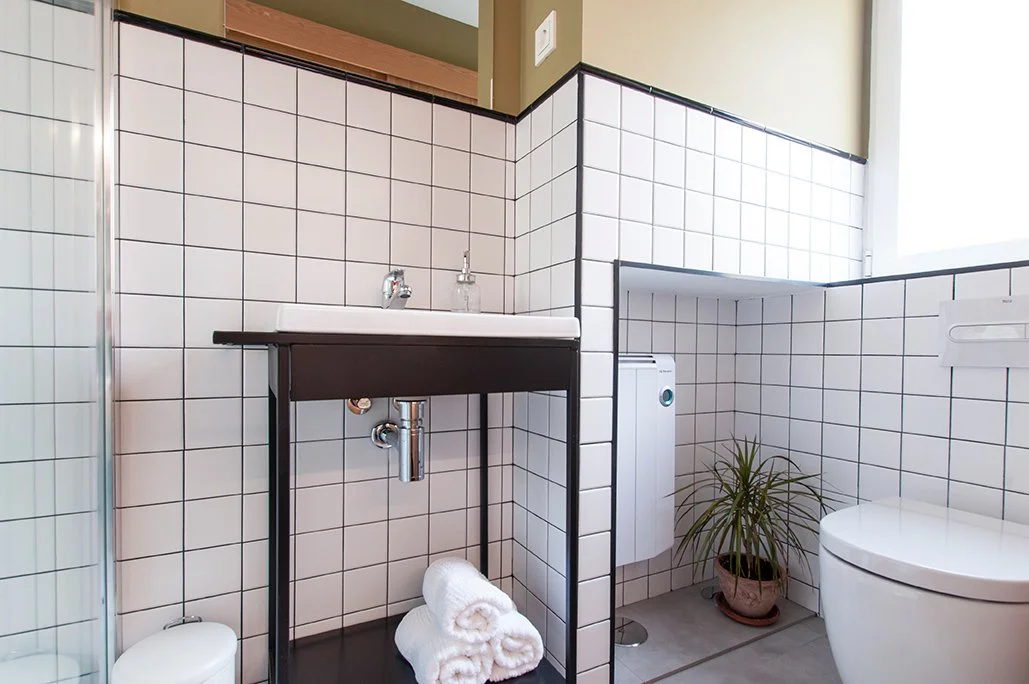 Baño moderno con azulejos blancos, lavabo con grifo y toallas, inodoro y planta en maceta.