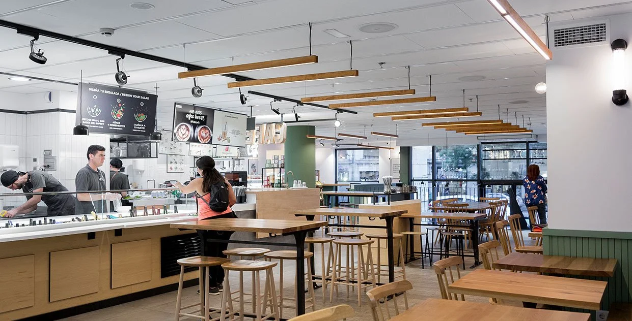 Un moderno restaurante o cafetería con estilo minimalista. Personas interactúan en el mostrador de comida. Hay mesas de madera y sillas en el área del comedor. Iluminación colgante y carteles en las paredes. Diseño de interiorismo comercial.