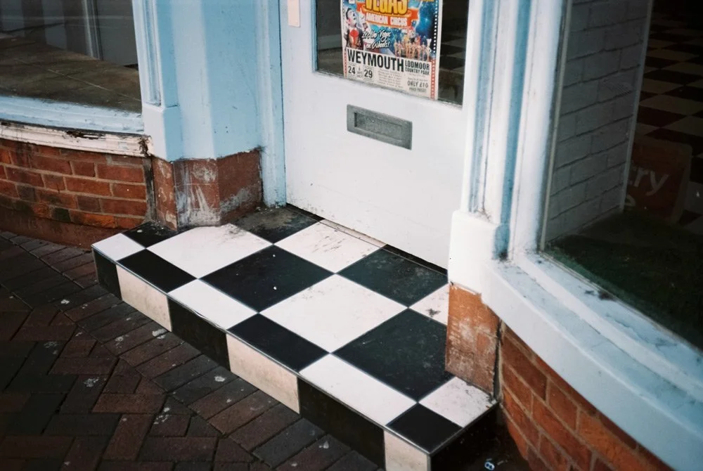 Entrada de tienda con piso de baldosas negras y blancas, cartel de anuncio en la puerta, y ventana con marco azul.