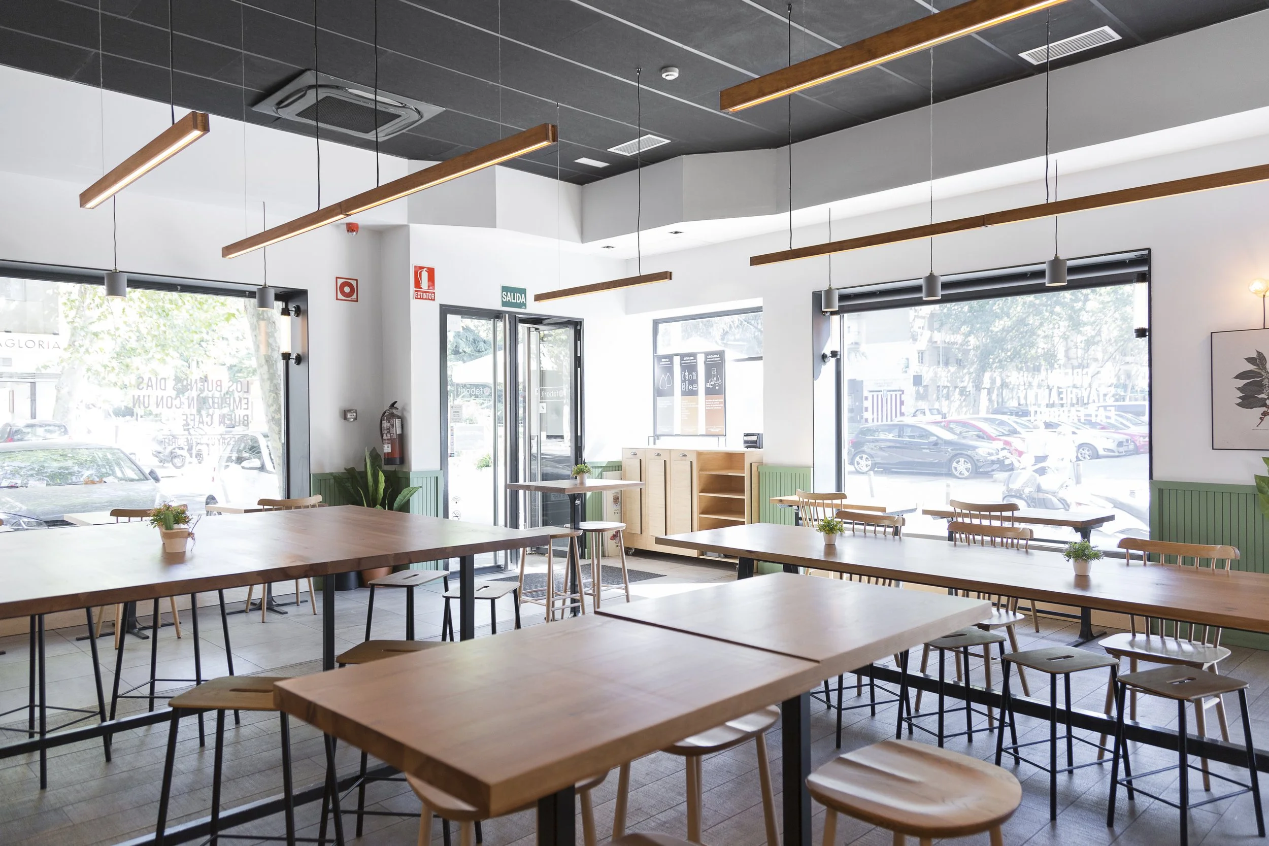 Interior de restaurante moderno con mesas de madera, sillas y luces colgantes, junto a grandes ventanales que ofrecen vista a la calle.