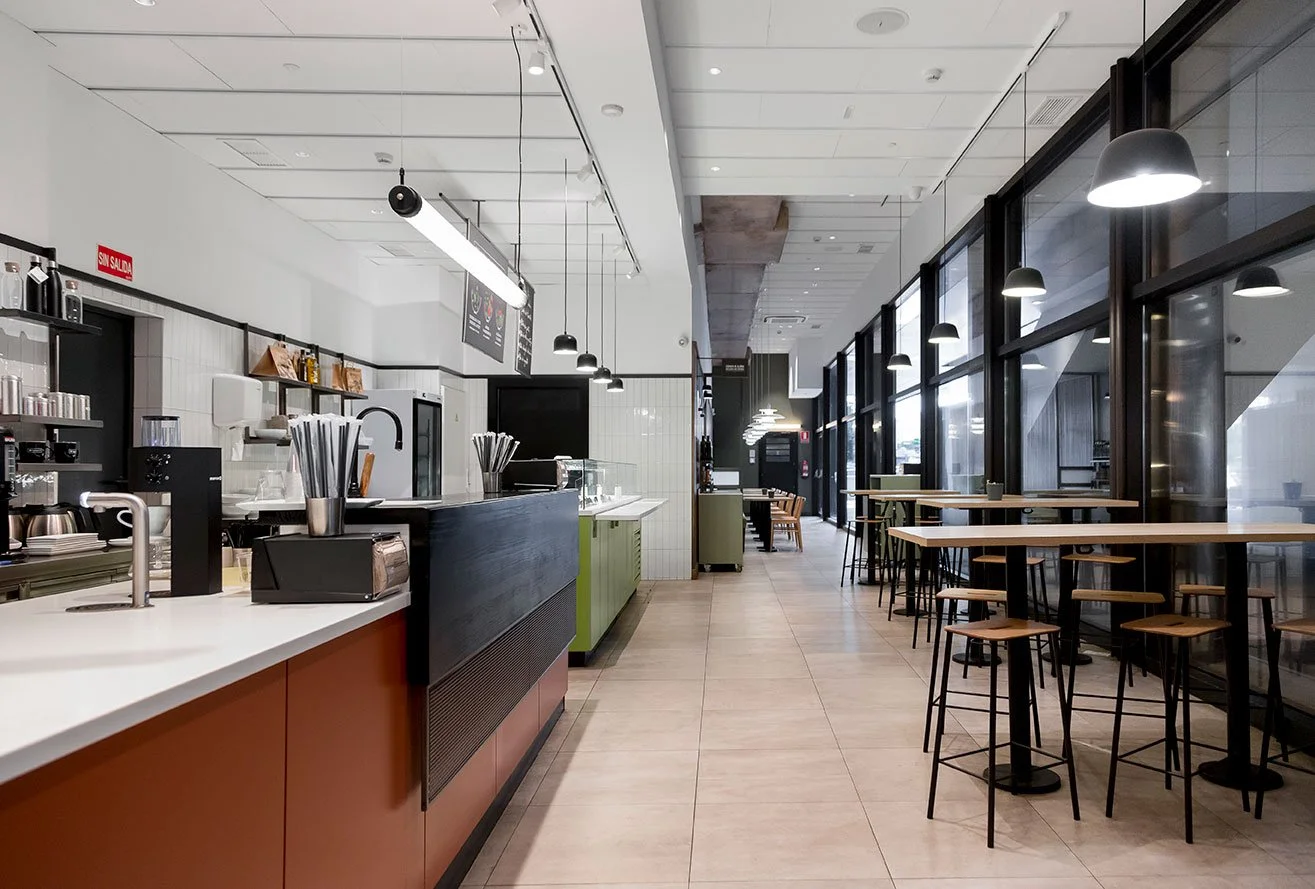 Cafetería moderna con mostrador, sillas altas y mesas altas, iluminación colgante y ventanas grandes. Diseño de interiorismo comercial. Retail design. Diseño de bares y restaurantes. Foto de Eva Carasol.