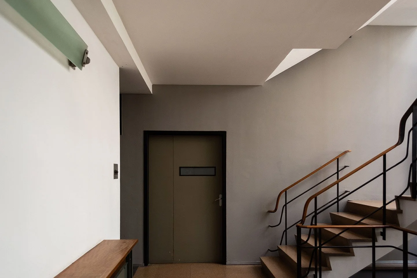 Interior de un edificio con escaleras de madera y barandales de metal, puerta de ascensor en la pared de fondo y pared blanca a la izquierda.