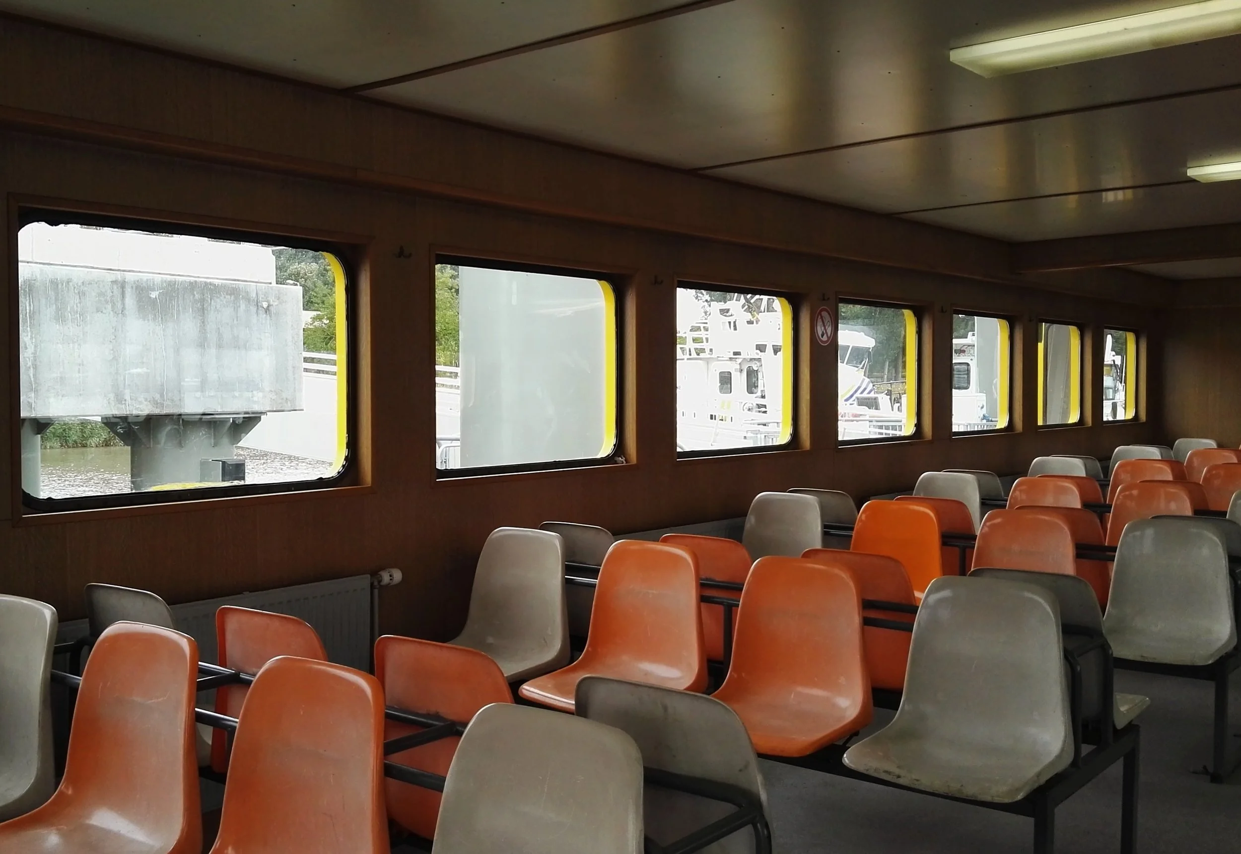 Interior de una sala en un ferry con asientos de color naranja y gris, y ventanas que muestran barcos y agua afuera. Antwerp.