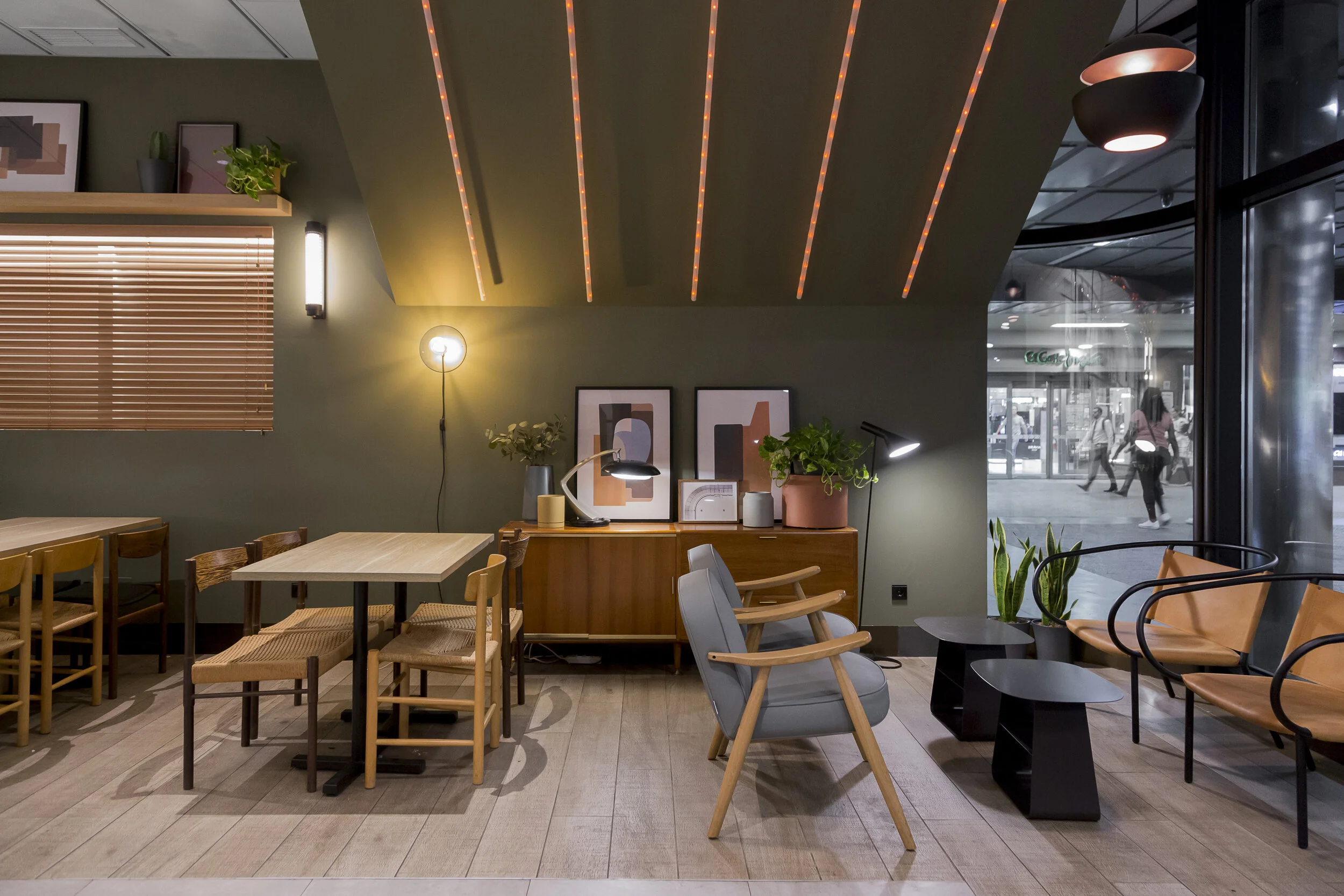 Interior de un café moderno con muebles de madera, sillas alrededor de mesas, plantas decorativas, lámparas de pared y techo, y una gran ventana que muestra el exterior. El diseño es minimalista y acogedor. Diseño de interiorismo comercial.