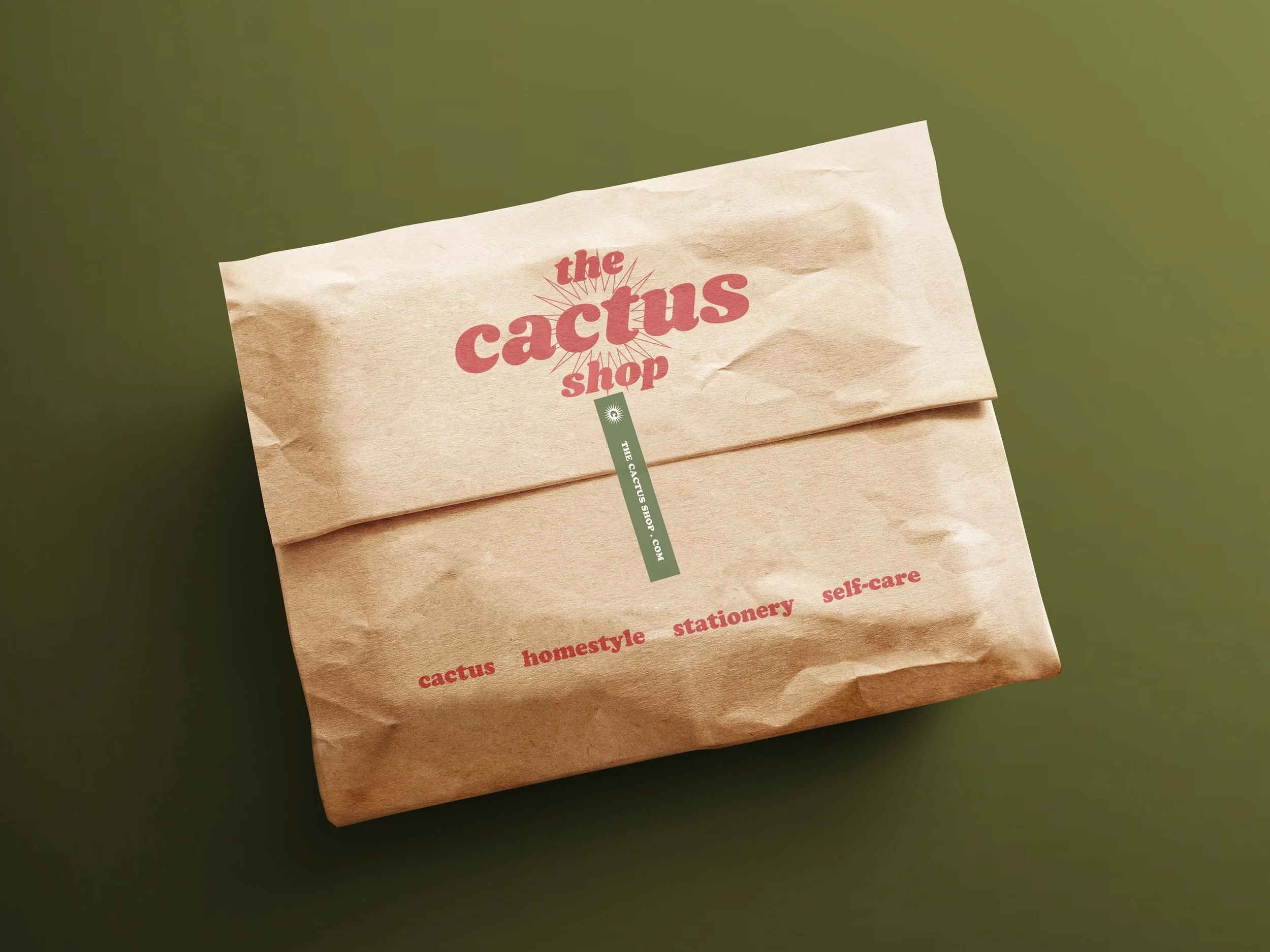 Bolsa de papel marrón con el logo de "The Cactus Shop", sobre un fondo verde. Letras impresas en rojo mencionan "cactus", "homestyle", "stationery", y "self-care". The Cactus Shop, diseño gráfico, branding y packaging. Agosto Creative Studio.