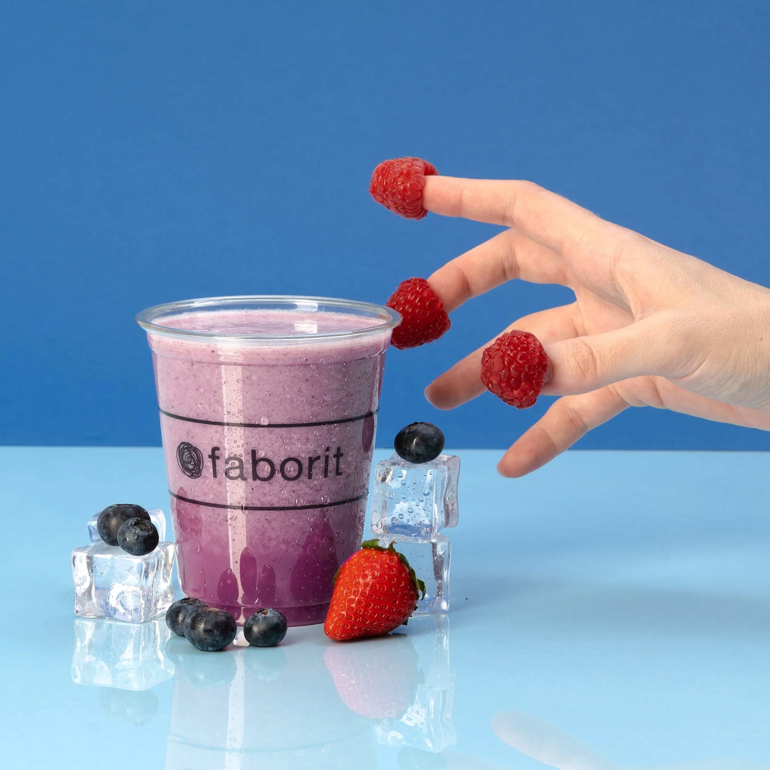Palillo con frambuesa en la punta, fruta en hielo, vaso con batido rosa, fresas, arándanos y hielo en una superficie azul.