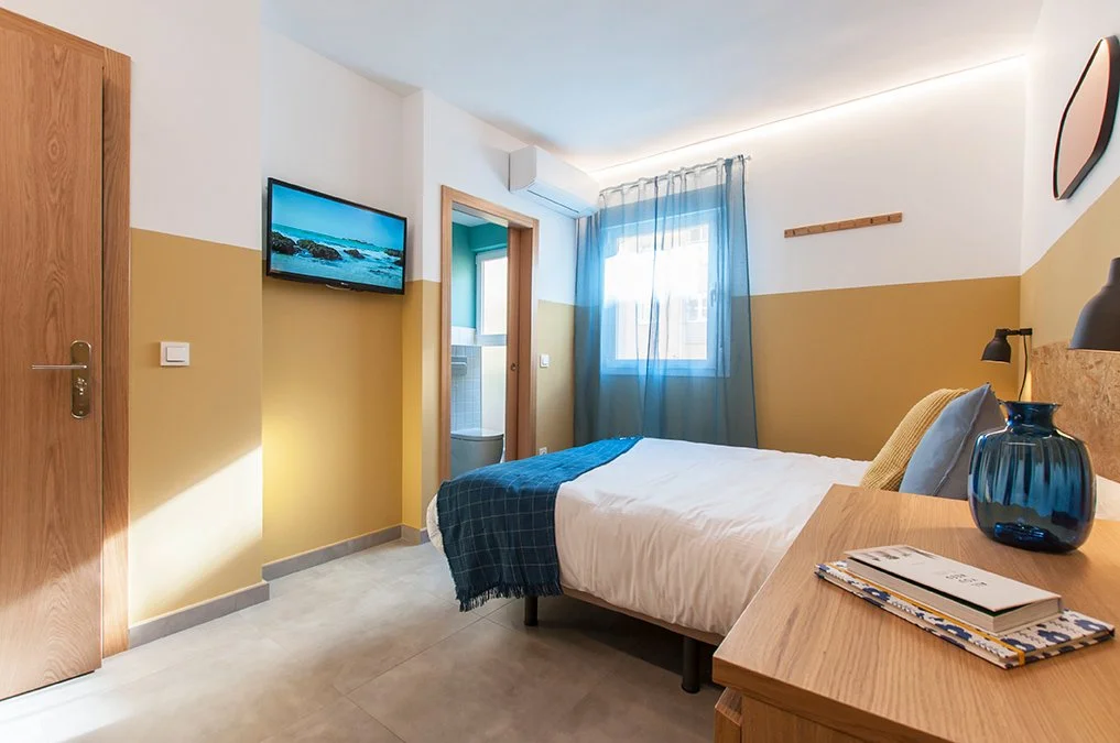 Habitación de hotel con cama individual, televisor en la pared, mesita de noche con libros, ventana con cortinas azules, lámpara, y puerta de baño abierta. Decoración moderna y minimalista.