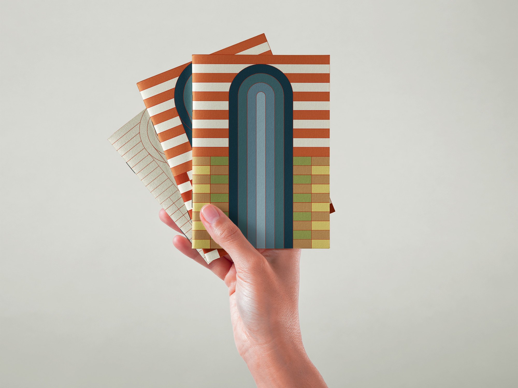 Mano sosteniendo tres cuadernos con diseños geométricos y coloridos inspirados en la arquitectura de Frank Lloyd Wright. Ilustración geométrica por Agosto Creative Studio Barcelona. Agencia de diseño y comunicación Barcelona.