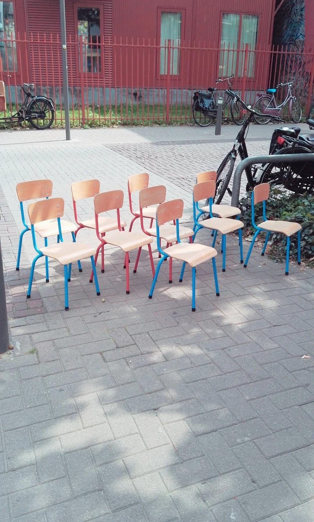 Sillas de colores en una mesa al aire libre, con bicicletas estacionadas en el fondo y un edificio rojo con ventanas.