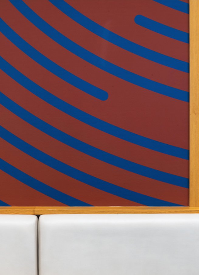 Panel decorativo con líneas curvas azules sobre fondo rojo, enmarcado con madera, con parte de un asiento blanco en la parte inferior.