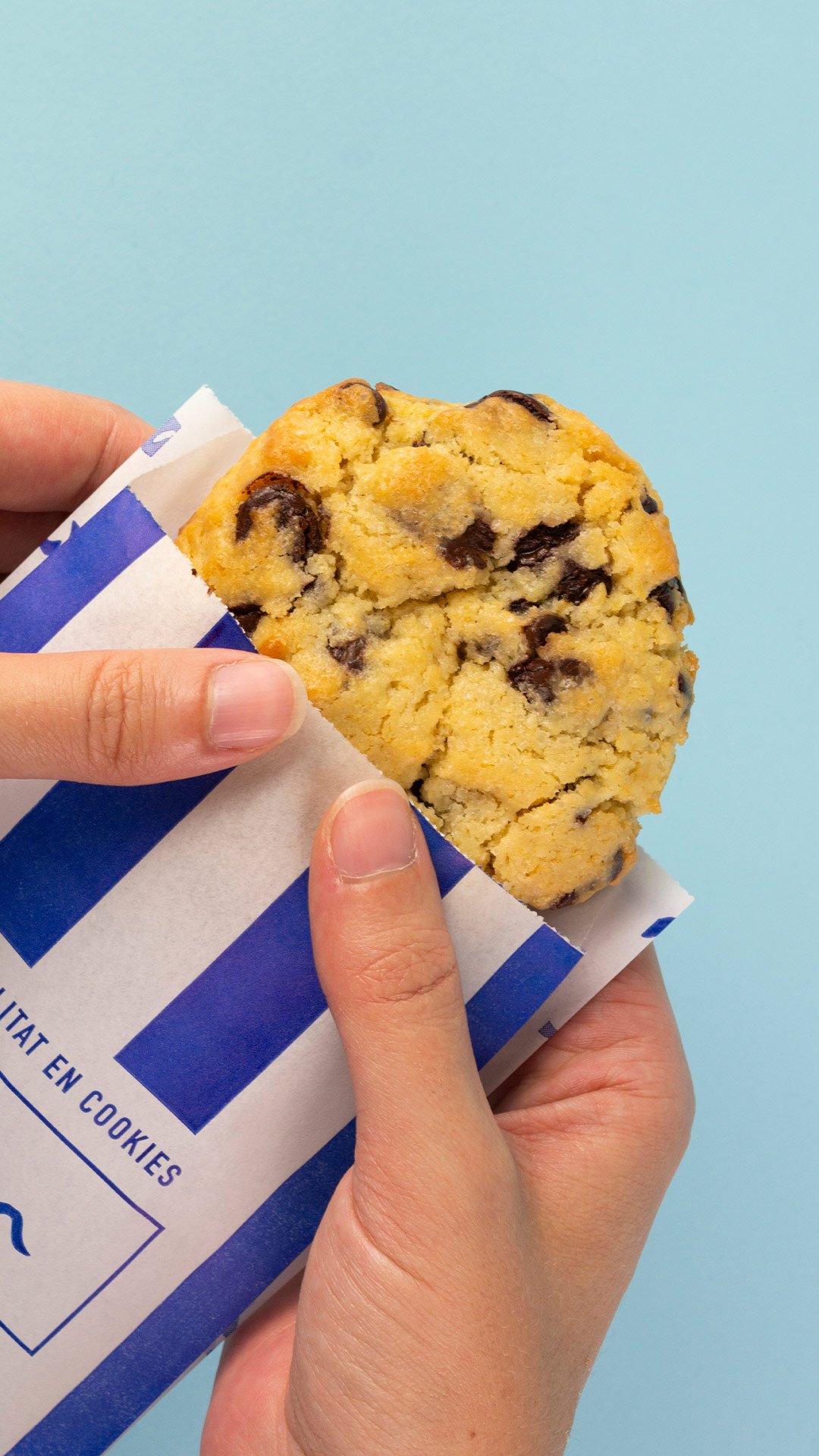 Galleta con chispas de chocolate en manos, en envoltura con rayas azules y blancas, fondo azul claro. Mionas Bakery. Especialidad en Cookies en Sitges. Retail Design, diseño de tiendas. Foto de producto.