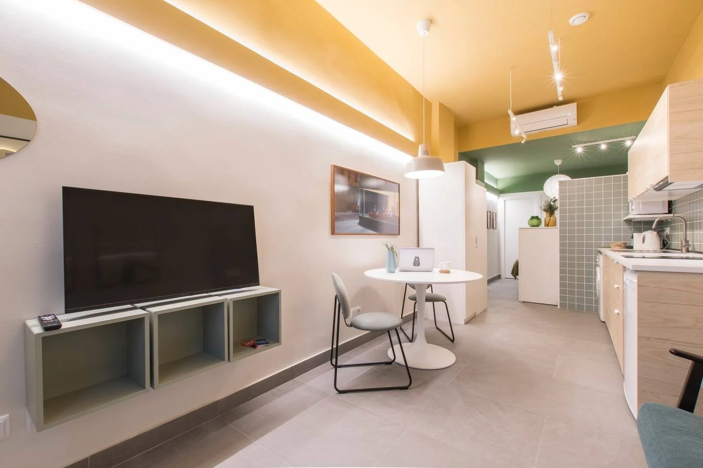 Sala de estar en un apartamento con televisión, mesa redonda, sillas, cocina y decoración moderna con colores pastel.