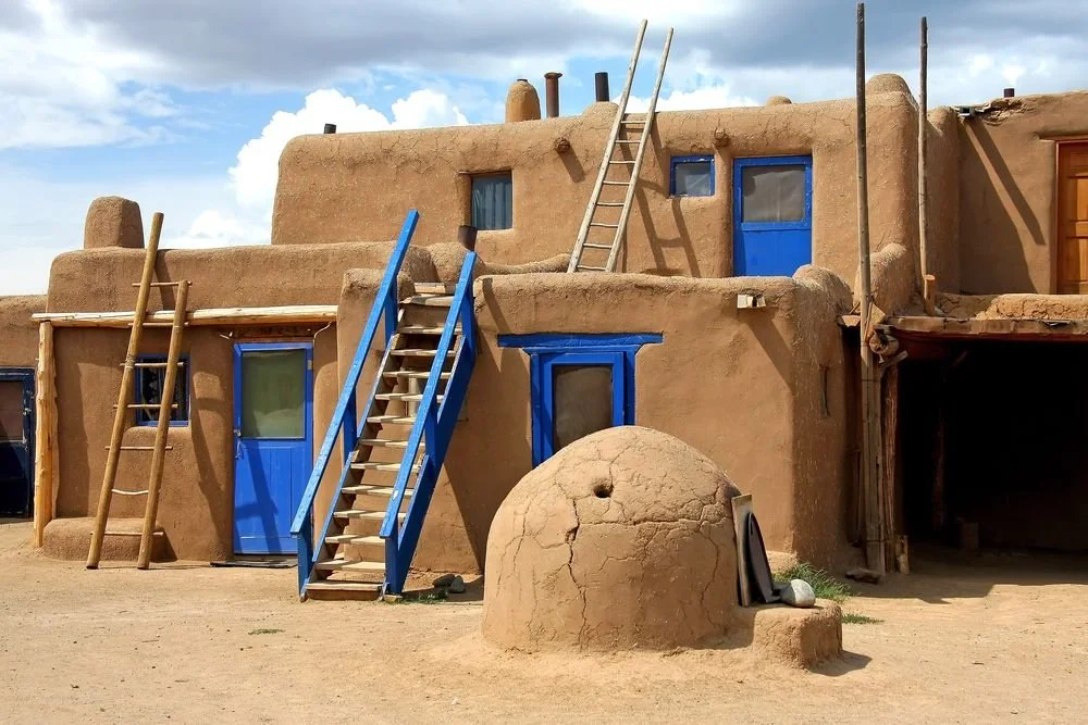 Casa de adobe tradicional con puertas y ventanas azules, escaleras de madera y horno de barro en el frente.