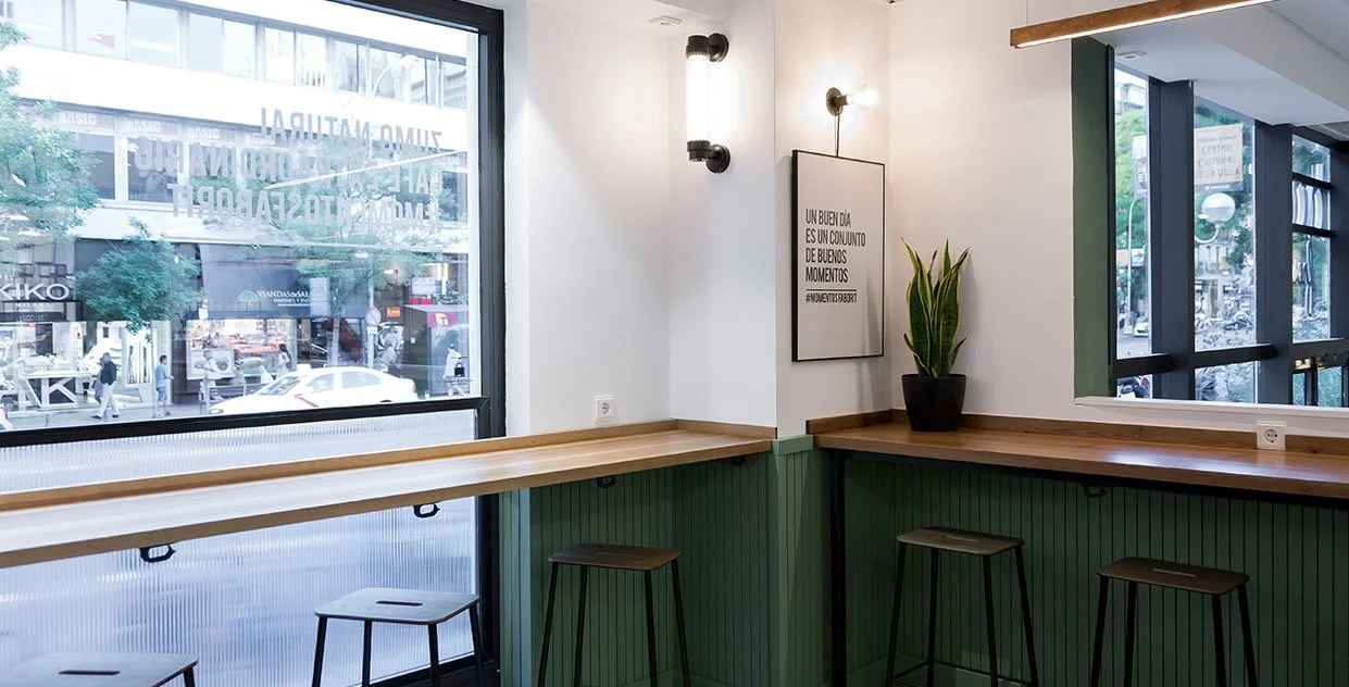 Interior de una cafetería moderna con mesas de madera, bancos altos y una planta en maceta. Un cartel en la pared dice 'Un buen día es un conjunto de buenos momentos'. Ventanas grandes permiten ver la calle y tiendas afuera. Diseño de interiorismo