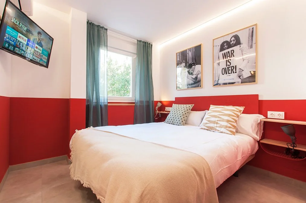 Habitación de hotel con cama doble, paredes rojas y blancas, televisor en la pared, ventana con cortinas verdes, y cuadros colgados sobre la cama.