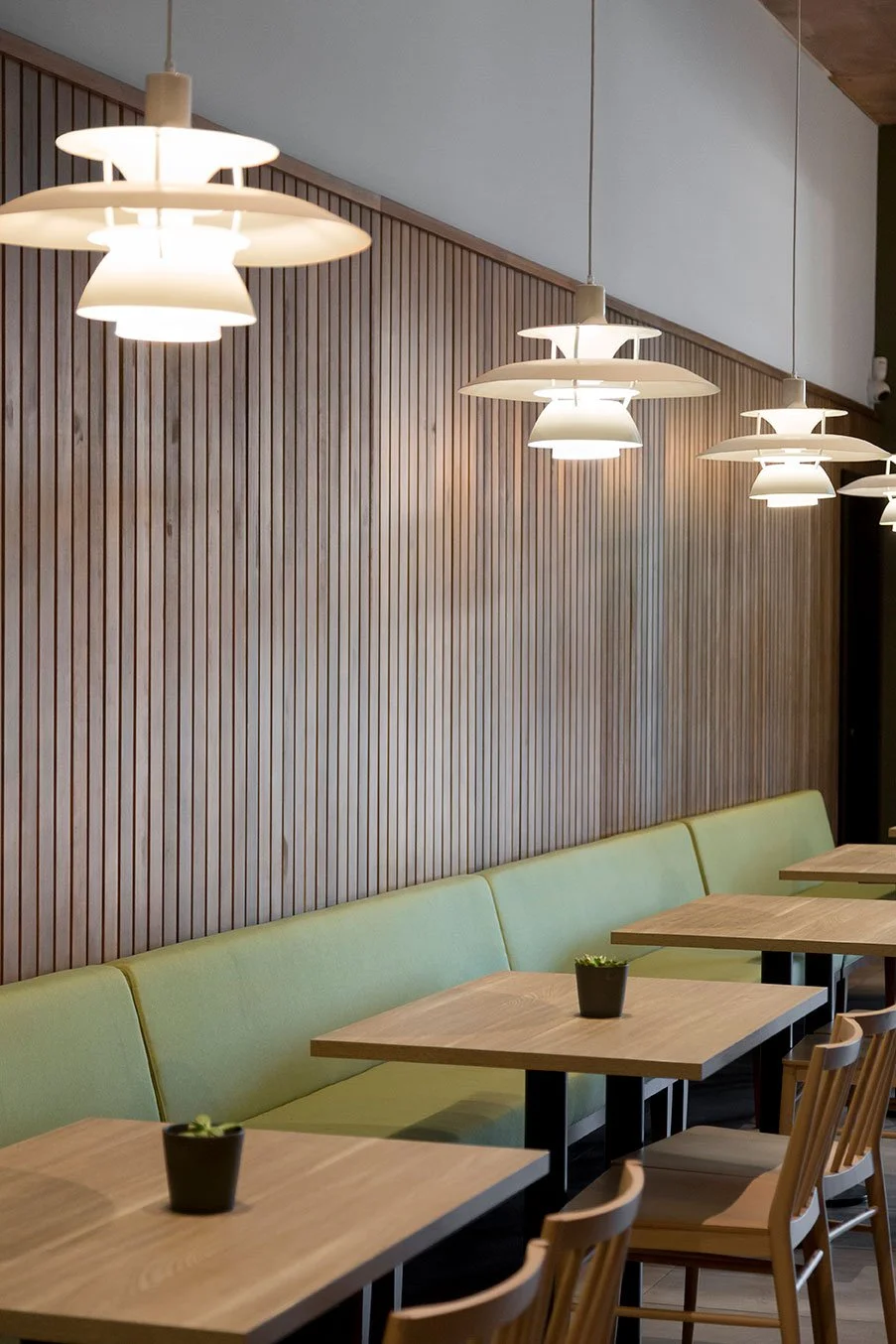 Interior de un café moderno con mesas de madera y sillas, lámparas colgantes y una pared con paneles de madera. Diseño de interiorismo comercial. Retail design. Diseño de bares y restaurantes. Foto de Eva Carasol.