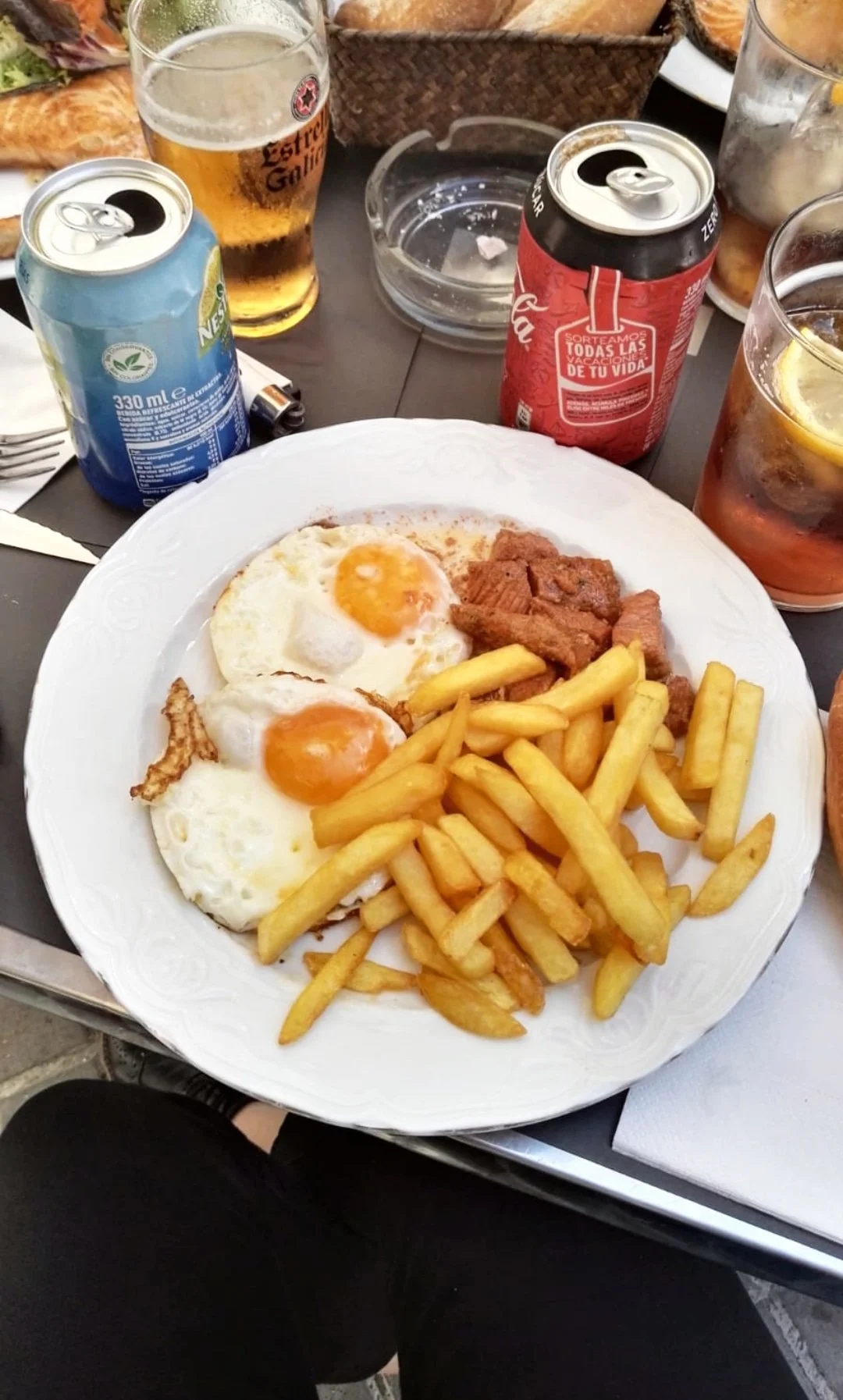 Plato con huevos estrellados, papas fritas y carne, rodeado de latas de bebidas, vasos con cerveza y refresco, en una mesa con comida adicional y utensilios.