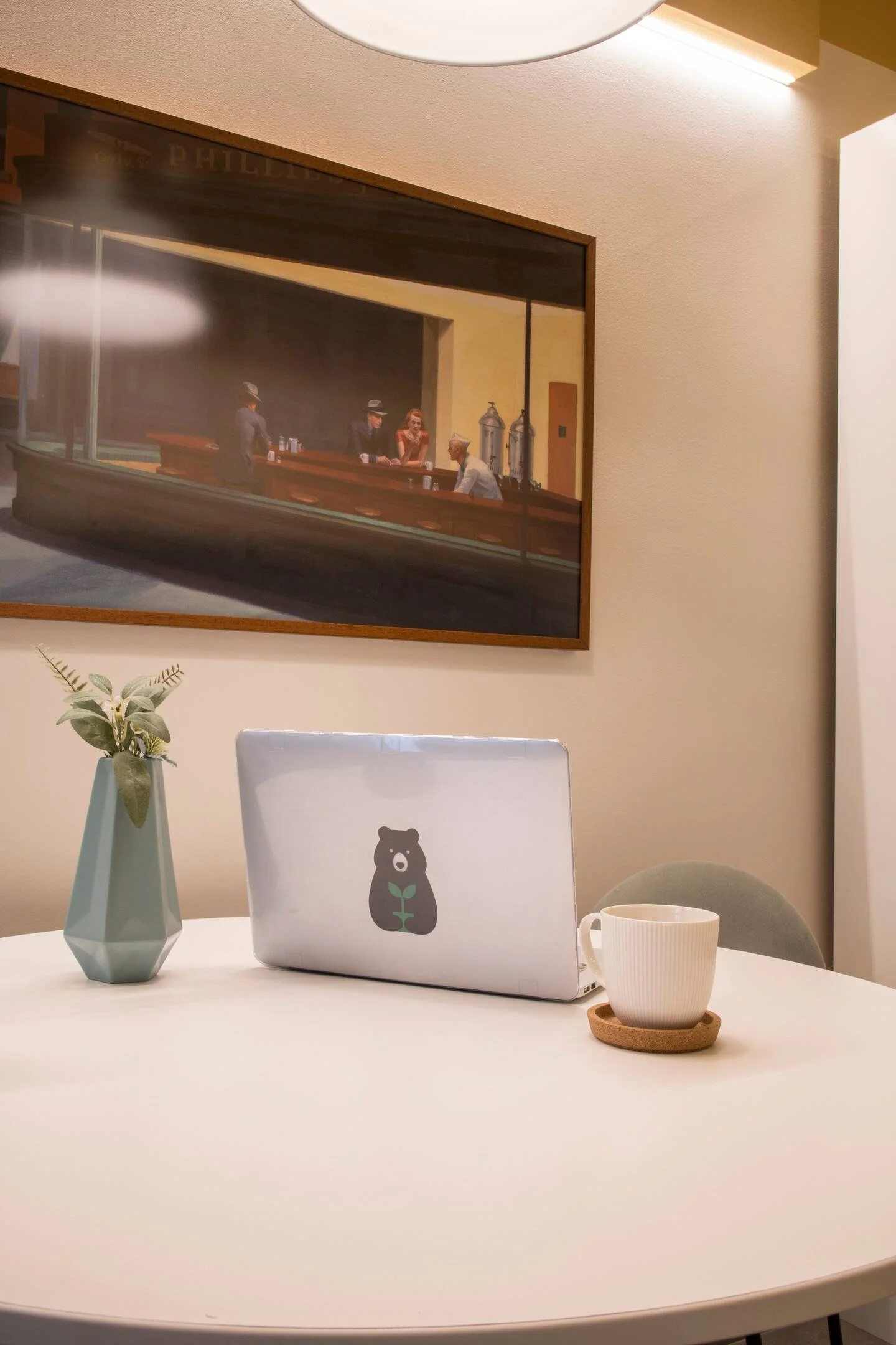 Mesa de trabajo con una laptop con sticker de oso y planta, una taza blanca con plato de corcho, una maceta con plantas, y acompañada de un cuadro con escena de bar en la pared.