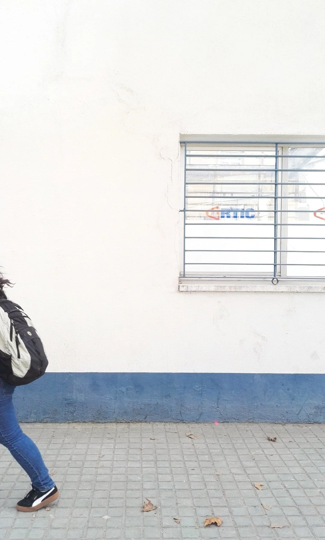 Una mujer caminando con mochila en una acera de concreto frente a una pared blanca con borde azul y una ventana con rejas.