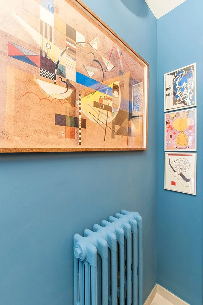 Esquina de una habitación con paredes azul cielo, radiador pintado de azul, y cuatro cuadros de arte abstracto colgados.