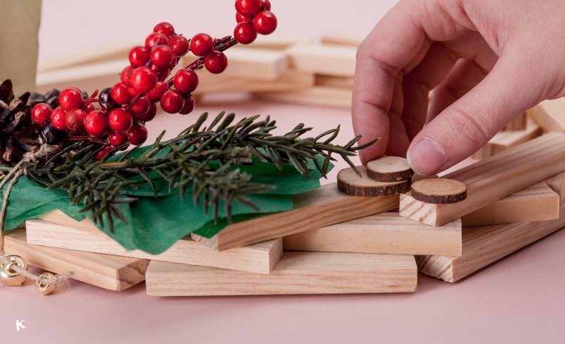 Persona colocando pequeñas piezas de madera en una estructura, con decoraciones navideñas, como ramas de pino y bayas rojas, en un fondo rosado.