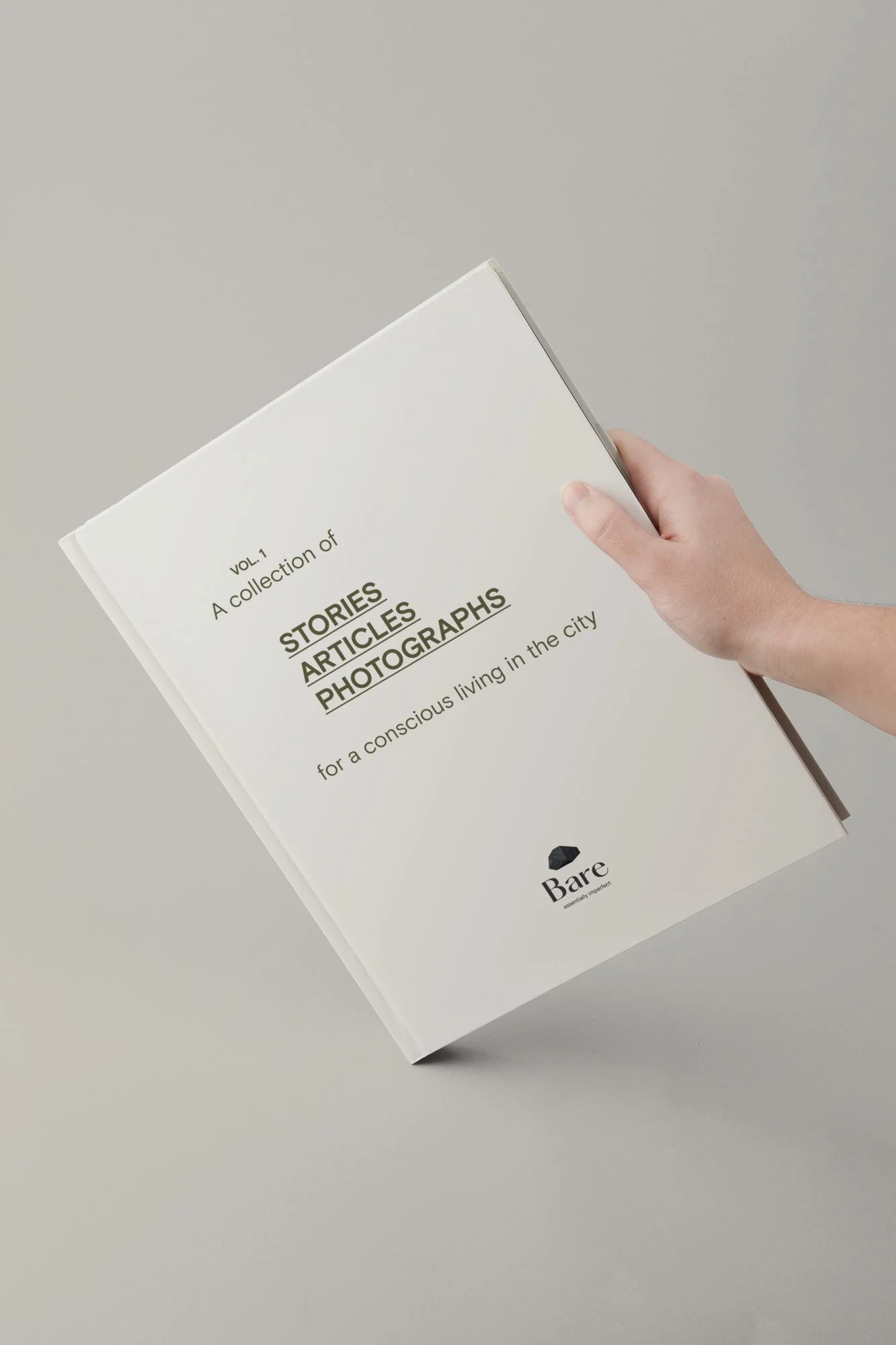 Mano sosteniendo un libro con texto en la portada que dice "A collection of stories, articles, photographs for a conscious living in the city." Marca "Bare" mencionada en la parte inferior.