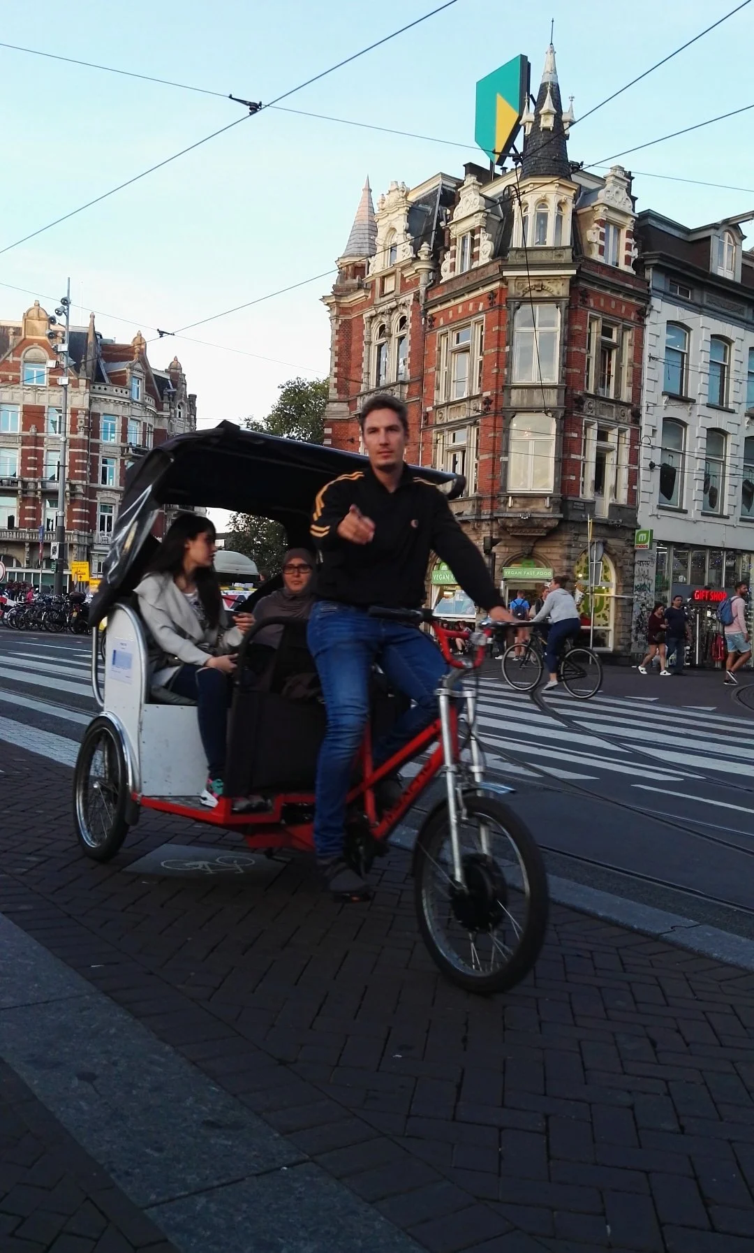 Un hombre conduce una bicicleta tándem con carruaje, acompañado de dos mujeres en la ciudad, con edificios históricos y líneas de tranvía en el fondo. Amsterdam.