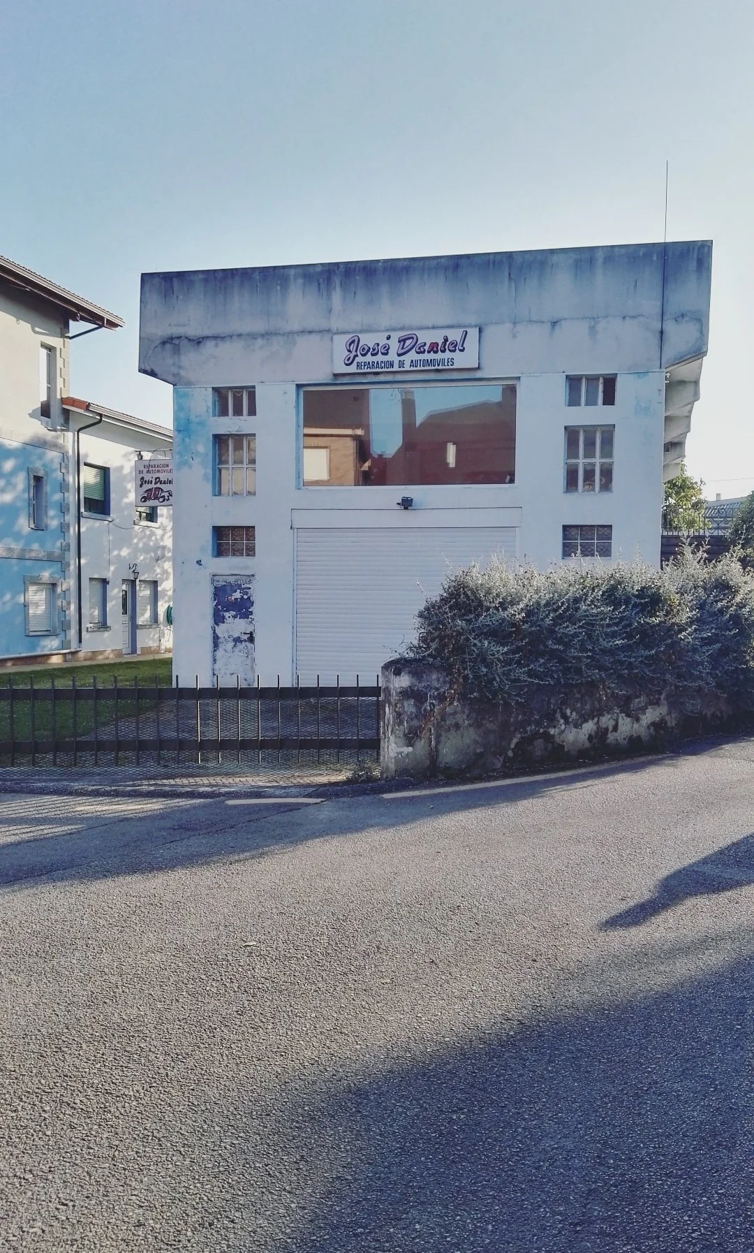 Edificio con cartel que dice 'José Daniel Reparación de Automóviles' en la calle, con un portón cerrado y vegetación en la acera del frente.