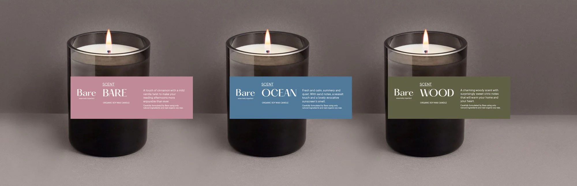 Tres velas aromáticas encendidas en frascos cilindricos negros, etiquetadas como Bare, Ocean y Wood, con descripciones de fragancias y cera de soya orgánica. Las etiquetas son de colores rosa, azul y verde, respectivamente.