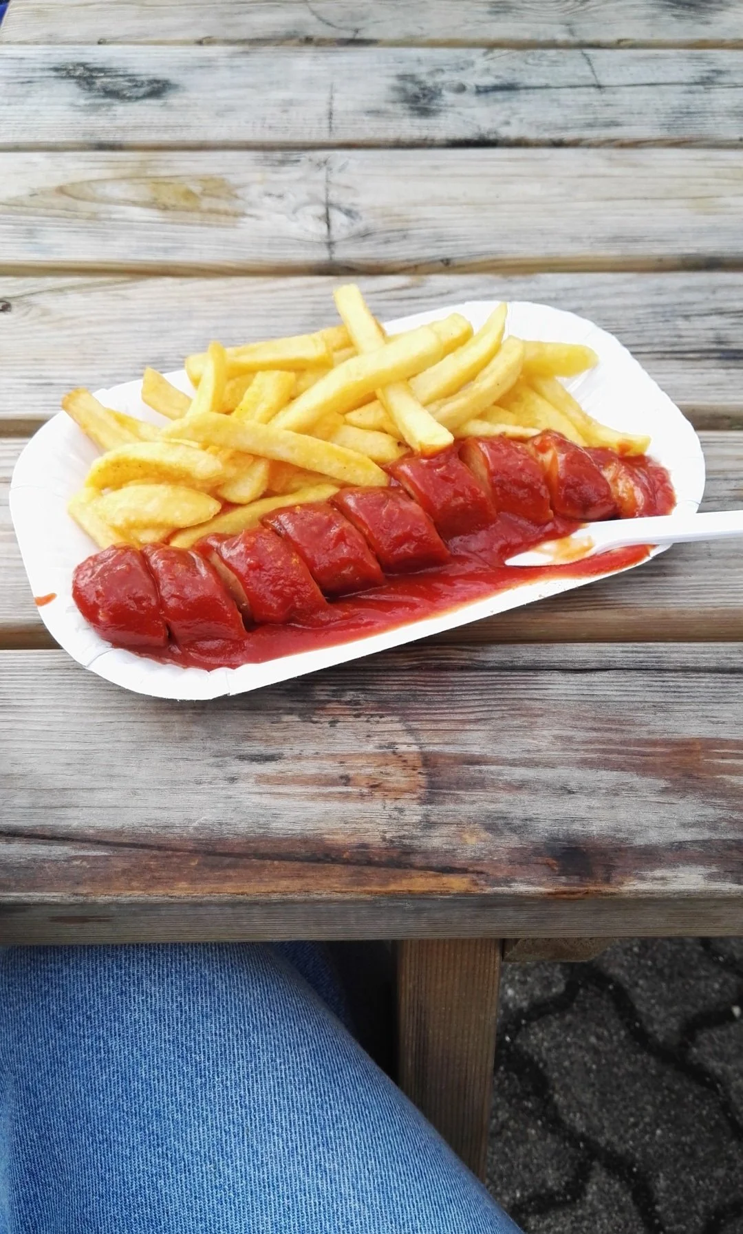Porción de papas fritas y una salchicha en salsa de tomate en un plato de papel sobre una mesa de madera. Currywurst.