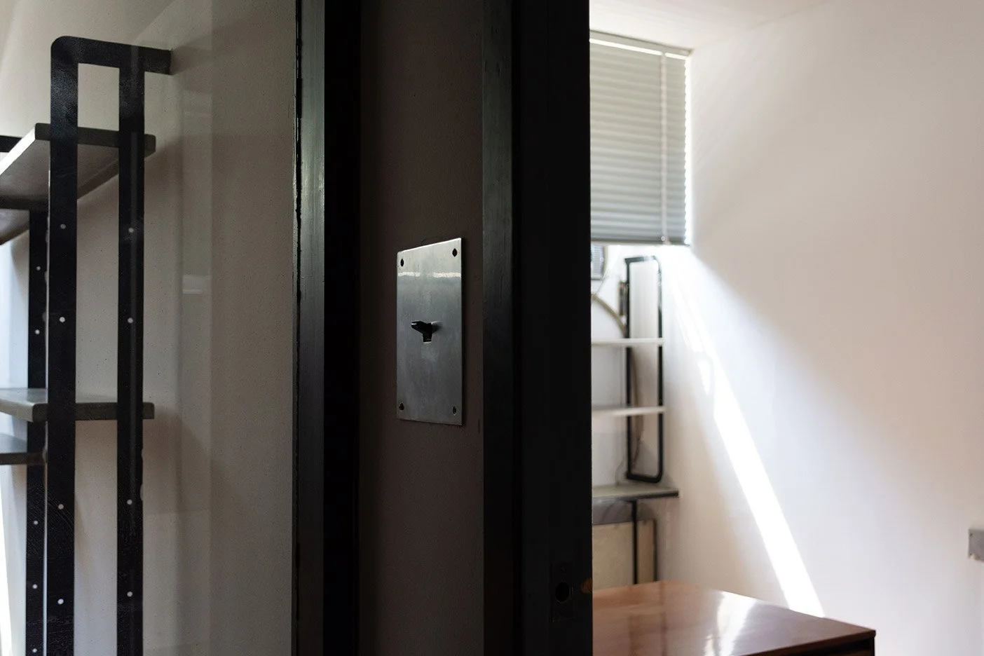 Vista de una habitación con una mesa de madera, una ventana con persiana y un espejo, y una estructura metálica negra en una esquina.