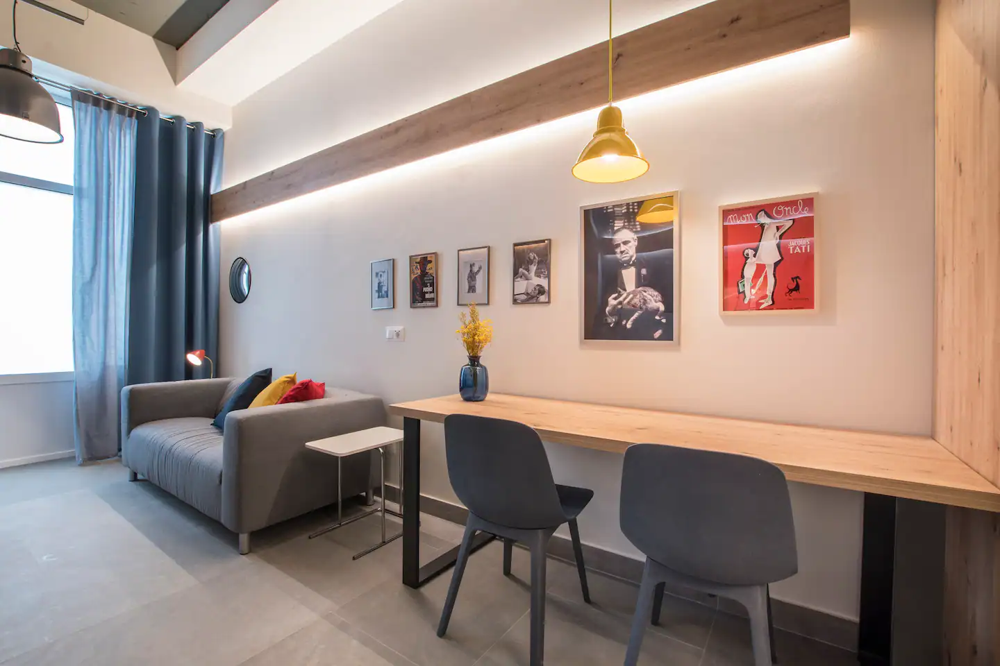 Sala de estar moderna con sofá gris, cortinas azules, mesa con vaso de flores, y pared decorada con fotografías y cuadros, iluminación tenue y ventana grande.