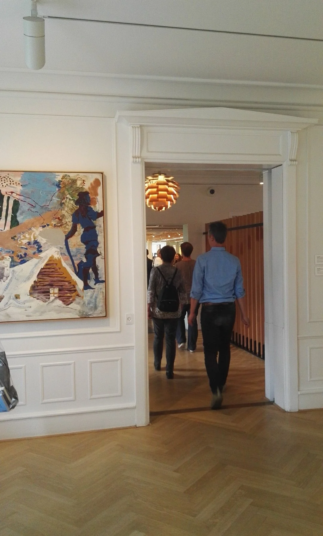 Algunos visitantes ingresan a una galería de arte o museo a través de una puerta decorada con molduras blancas, viendo un mural abstracto de colores vibrantes en la pared izquierda y una lámpara de diseño moderno en el techo. Louisiana Museum, Denmar