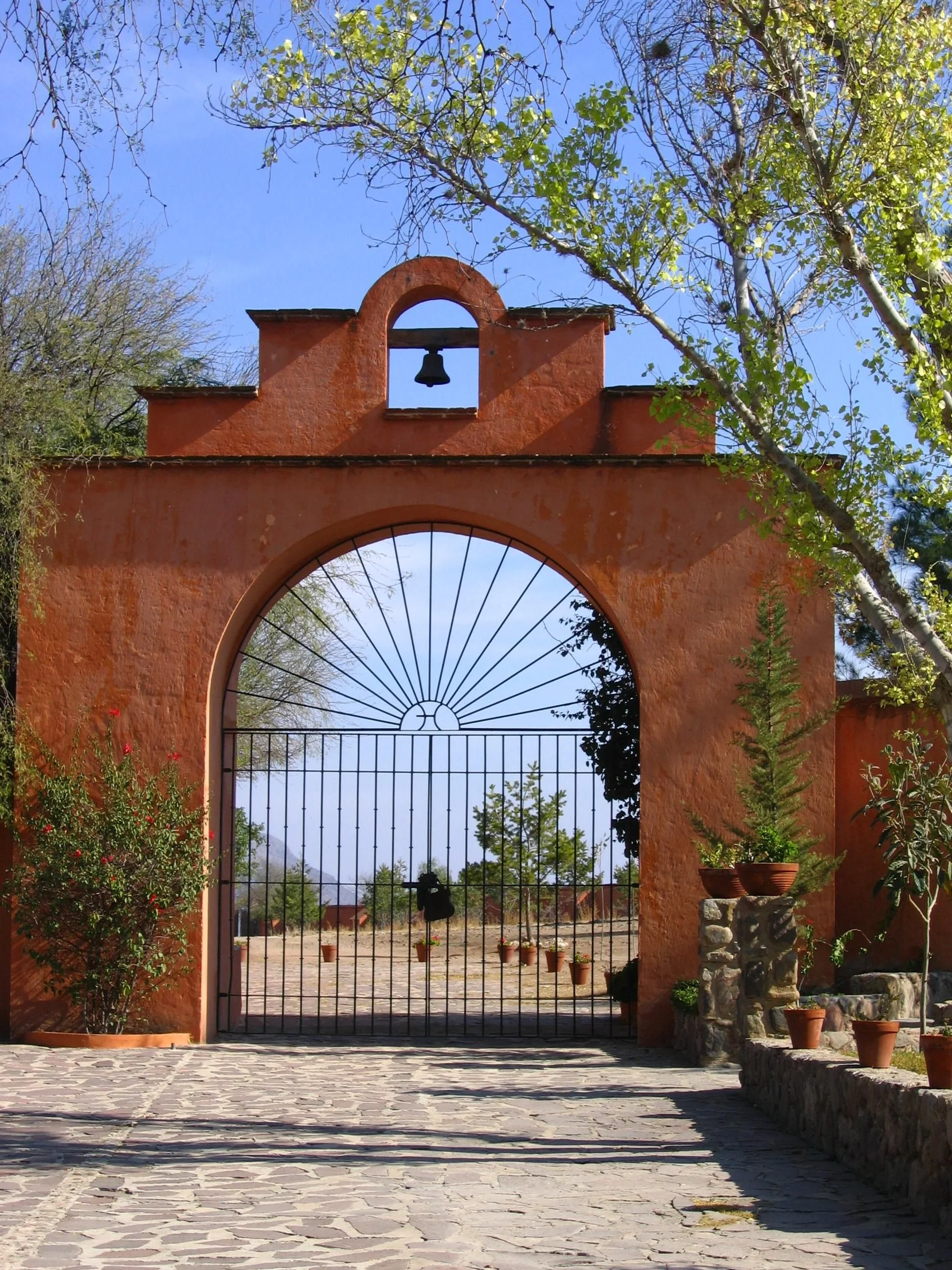 Entrada de arco con portón de hierro y campana en la parte superior, rodeada de árboles y plantas en macetas, bajo un cielo azul.