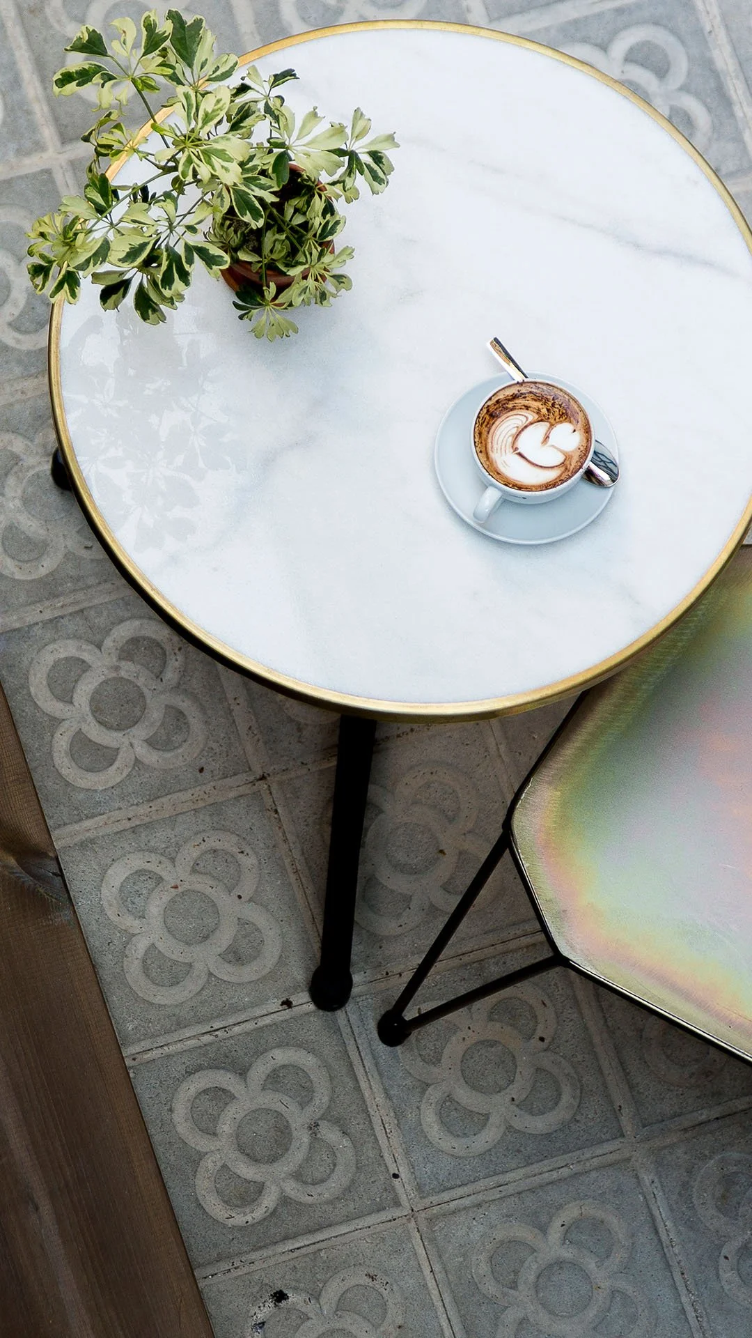Mesa redonda con superficie de mármol, con una planta en maceta y una taza de café con leche y arte latte, sobre un piso con azulejo con diseño floral.