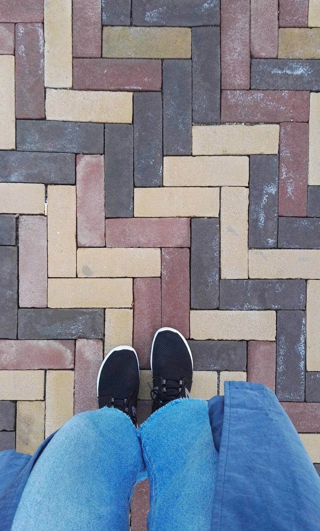Pavimento de bloques de piedra en patrón entrecruzado, con zapatos deportivos negros y pantalones azules en primer plano. Amsterdam floor tiles.