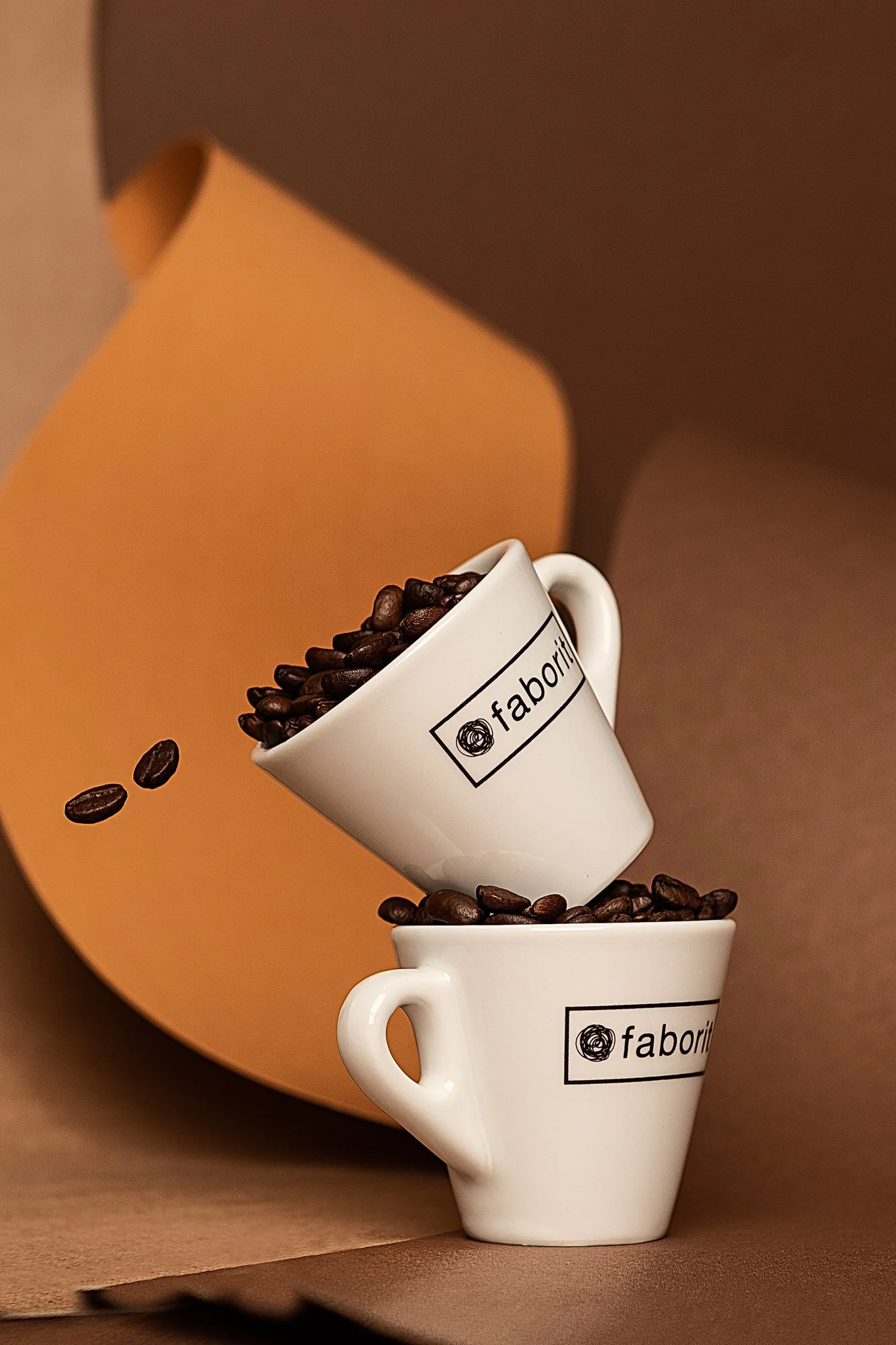 Dos tazas blancas con logo 'faborit' apiladas, llenas de granos de café, en un fondo marrón con una estructura decorativa
