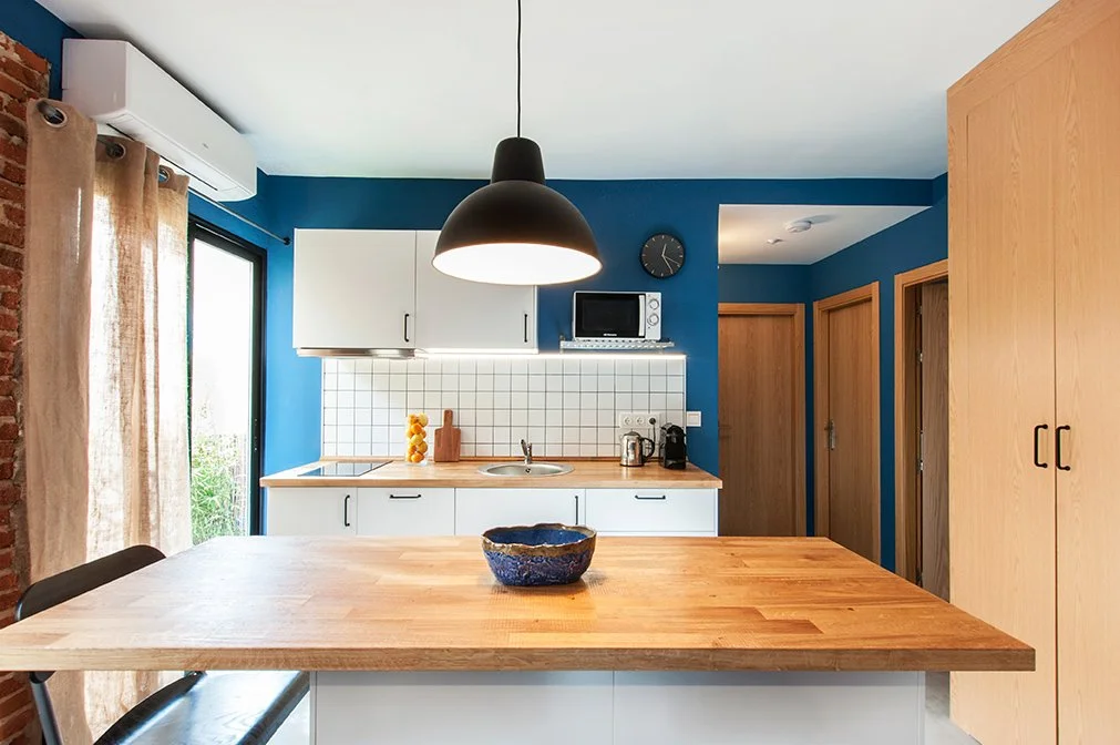 Cocina moderna con paredes azules, encimera de madera, lámpara colgante negra, ventana con cortinas beige, aire acondicionado, azulejos blancos, y electrodomésticos como microondas y cafetera. Hay un bol azul sobre la isla de cocina.