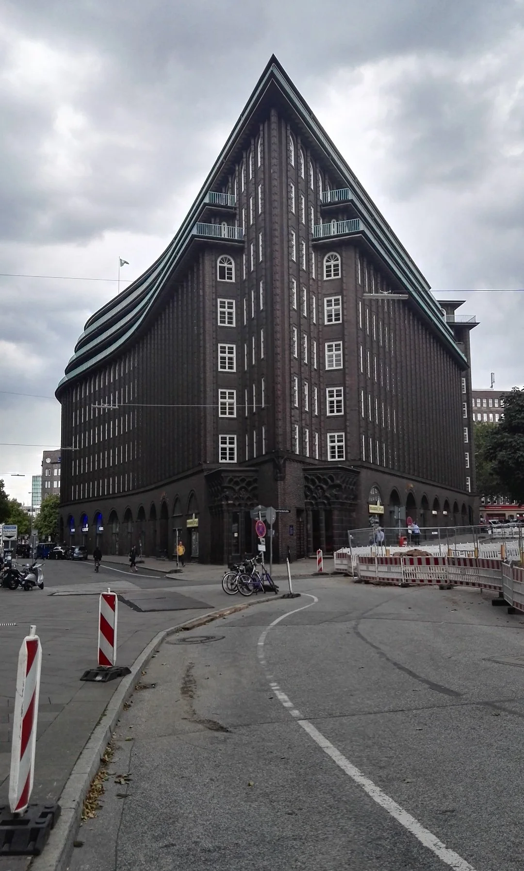 Edificio oscuro de varios pisos con ventanas blancas, ubicado en una esquina de la ciudad con calles en reparación, ciclistas y peatones, bajo un cielo nublado. Hamburg.