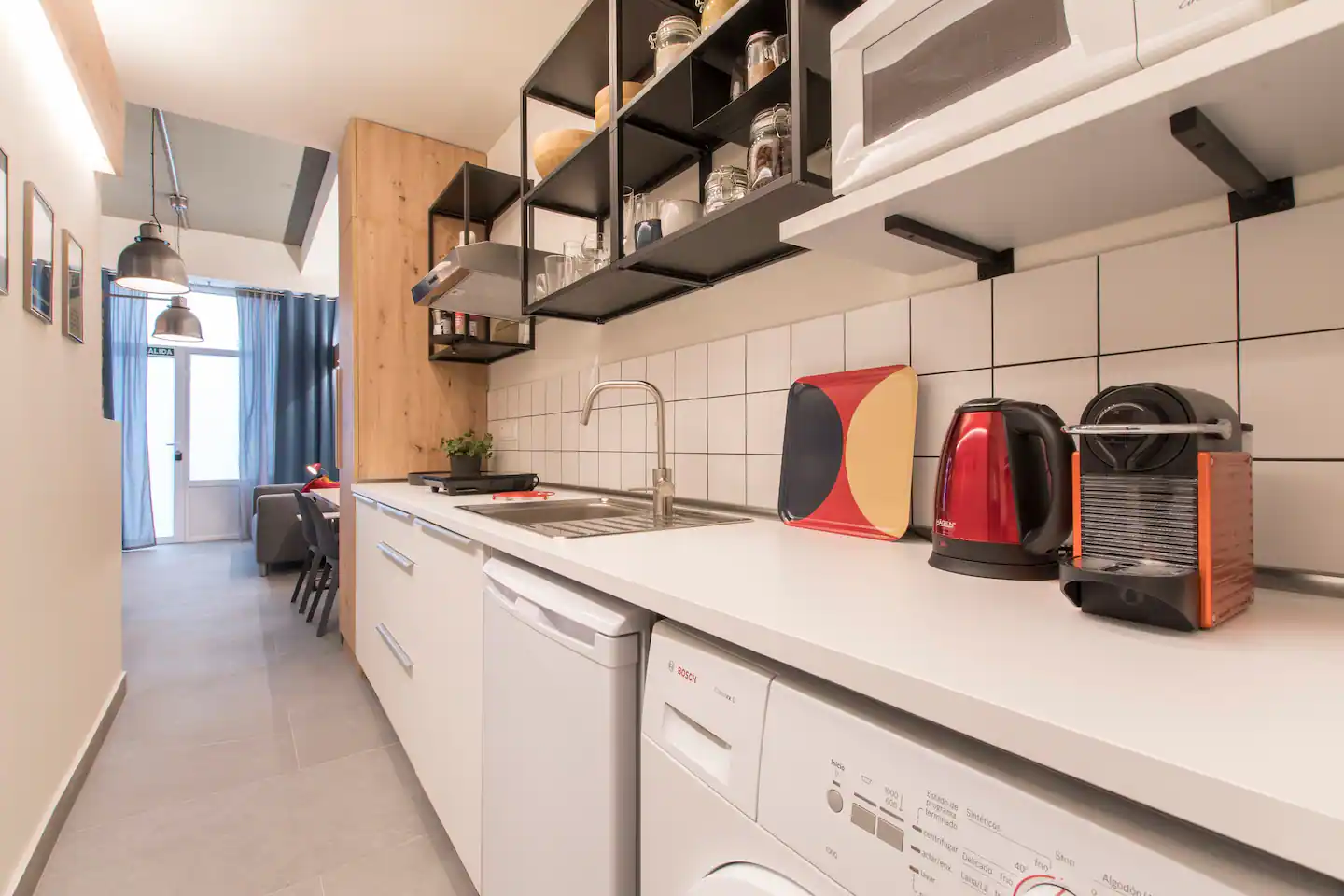 Cocina moderna con utensilios y electrodomésticos sobre la encimera y estanterías abiertas con utensilios y cristalería. Al fondo, un espacio de sala con sillas y ventanas con cortinas.