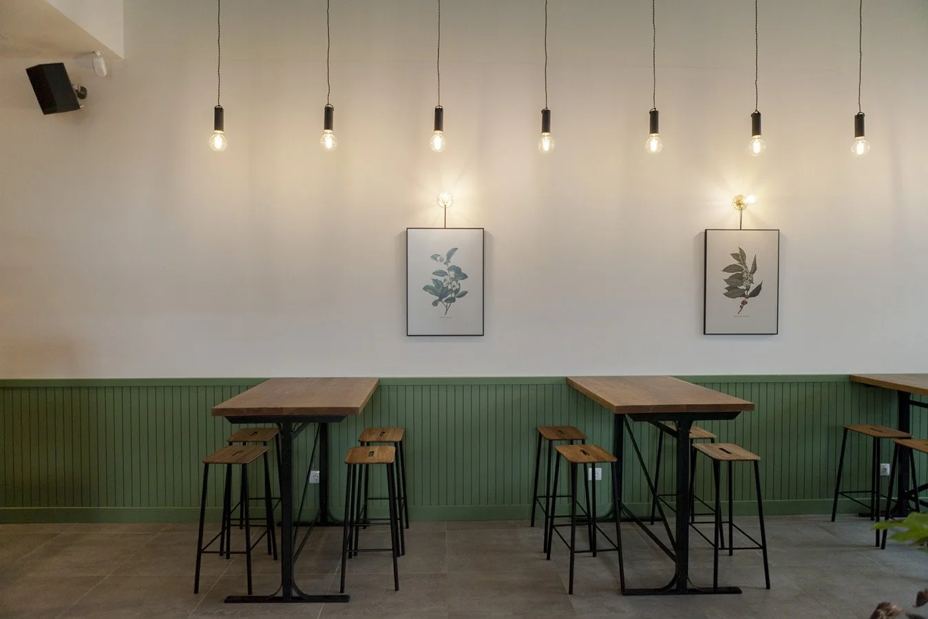 Interior de un café moderno con mesas altas de madera, taburetes y una pared decorada con cuadros botánicos y lámparas colgantes. Diseño de interiorismo comercial. Retail Design.