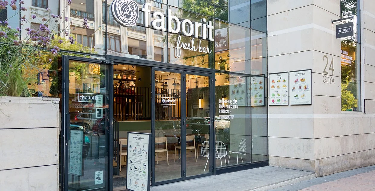 Entrada de una cafetería "faborit fresh bar" con ventanas de vidrio, mesas y sillas en el interior, ubicada en el número 24 de la calle Goya. Diseño de interiorismo comercial. Retail Design. Foto de Eva Carasol.