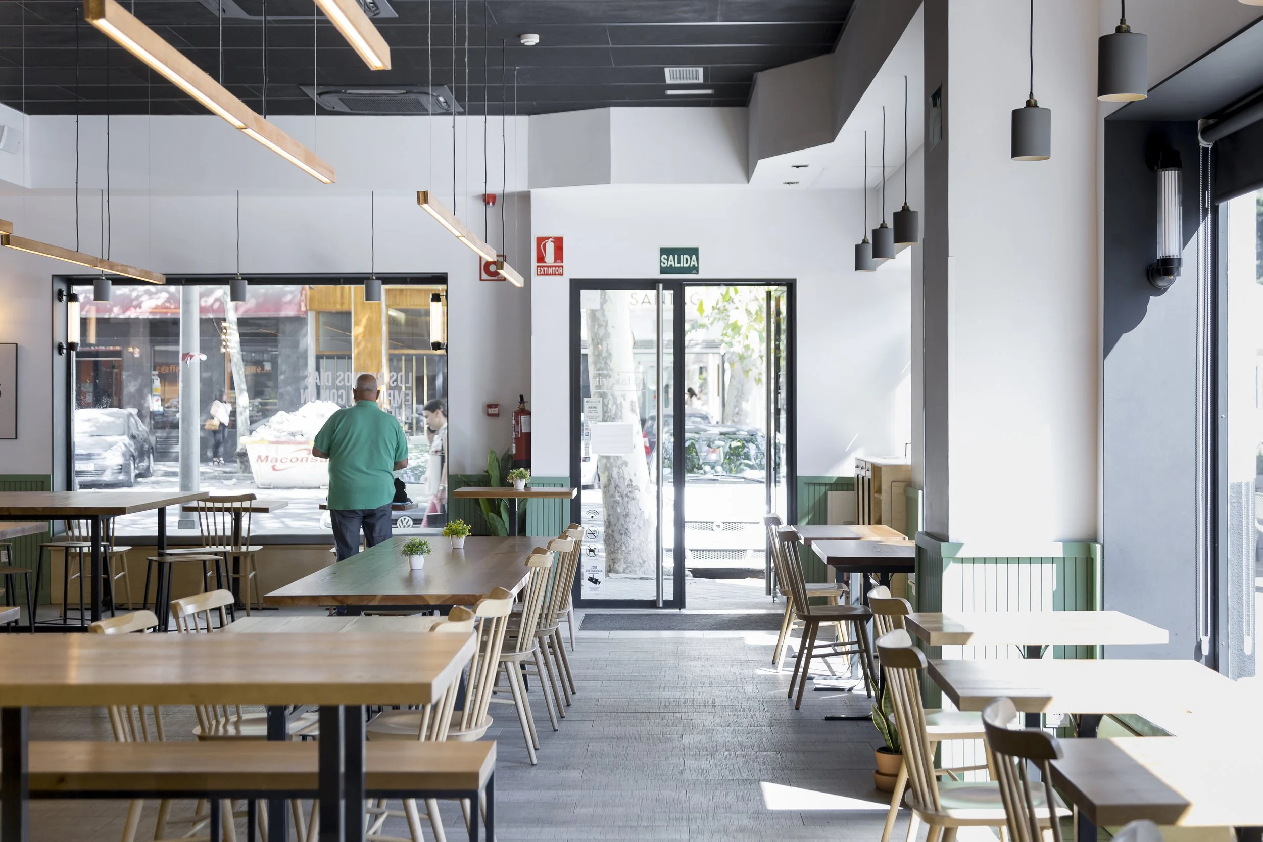 Interior de un restaurante moderno y luminoso con mesas y sillas de madera, lámparas colgantes y una salida con puerta de cristal. Una persona está de pie cerca de la ventana.