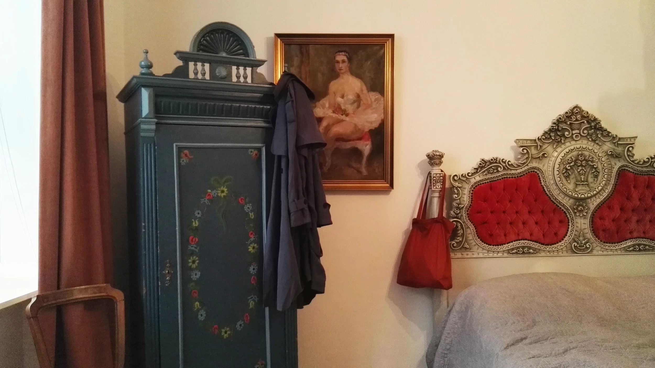 Habitación con armario azul con flores pintadas, pintura de mujer en el centro, cabecera de cama con detalles ornamentales y tapizado en rojo, colcha beige, cortinas marrones.