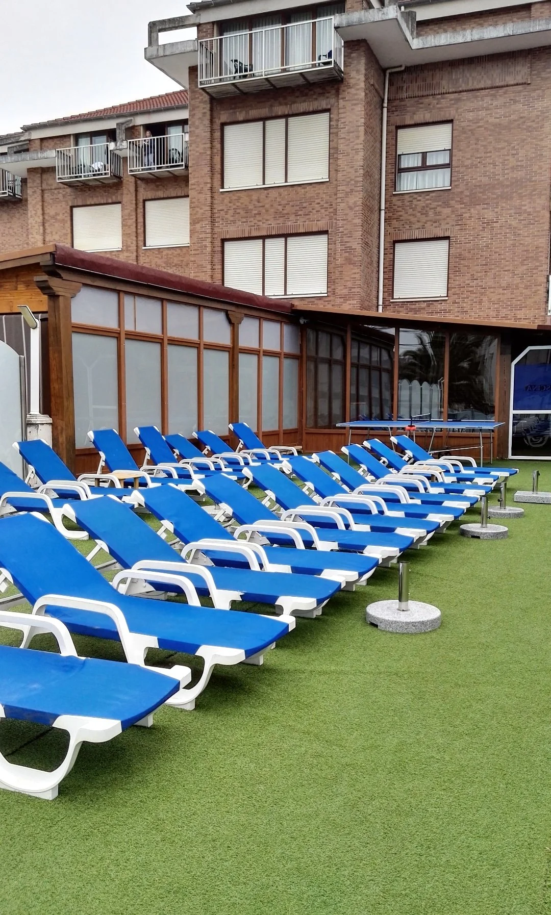 Una fila de tumbonas azules en una piscina, con una estructura de madera y un edificio de ladrillo al fondo.