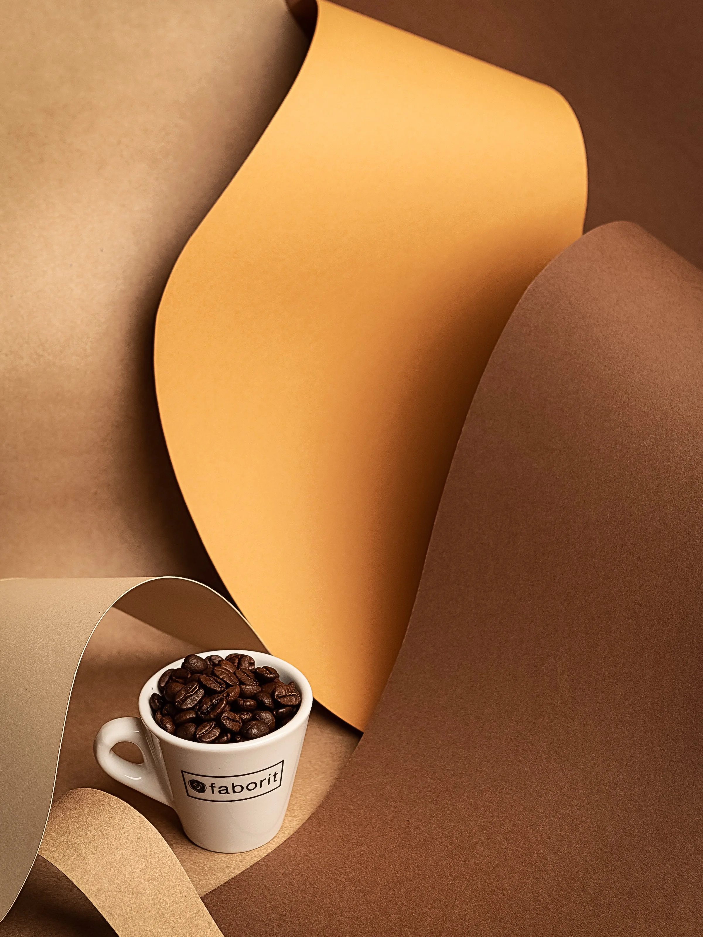 Taza blanca con granos de café sobre papel de color beige y marrón en un fondo de papeles de colores beige, marrón y naranja.