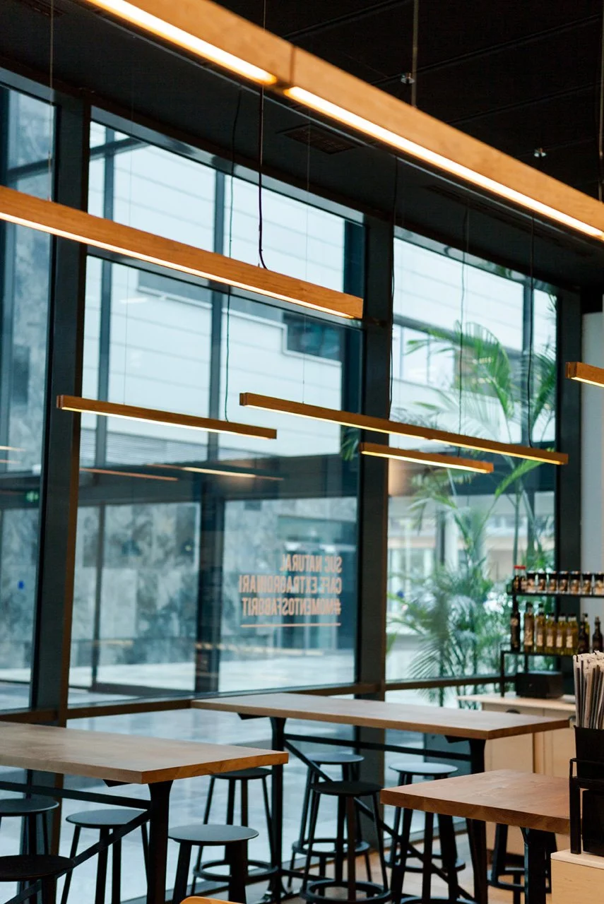 Interior de una cafetería moderna con mesas de madera, sillas altas y lámparas colgantes. Ventanales grandes y botellas en estantes al fondo.