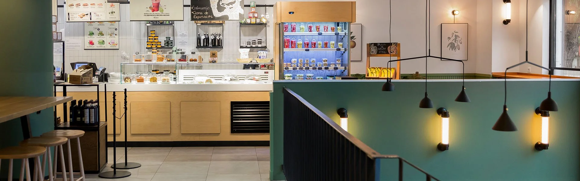 Interior de una cafetería moderna, con mostrador de venta de bebidas y alimentos frescos, estantes con productos y refrigerador lleno de botellas y envases. Mesas y sillas están dispuestas cerca de las paredes decoradas con luces colgantes.