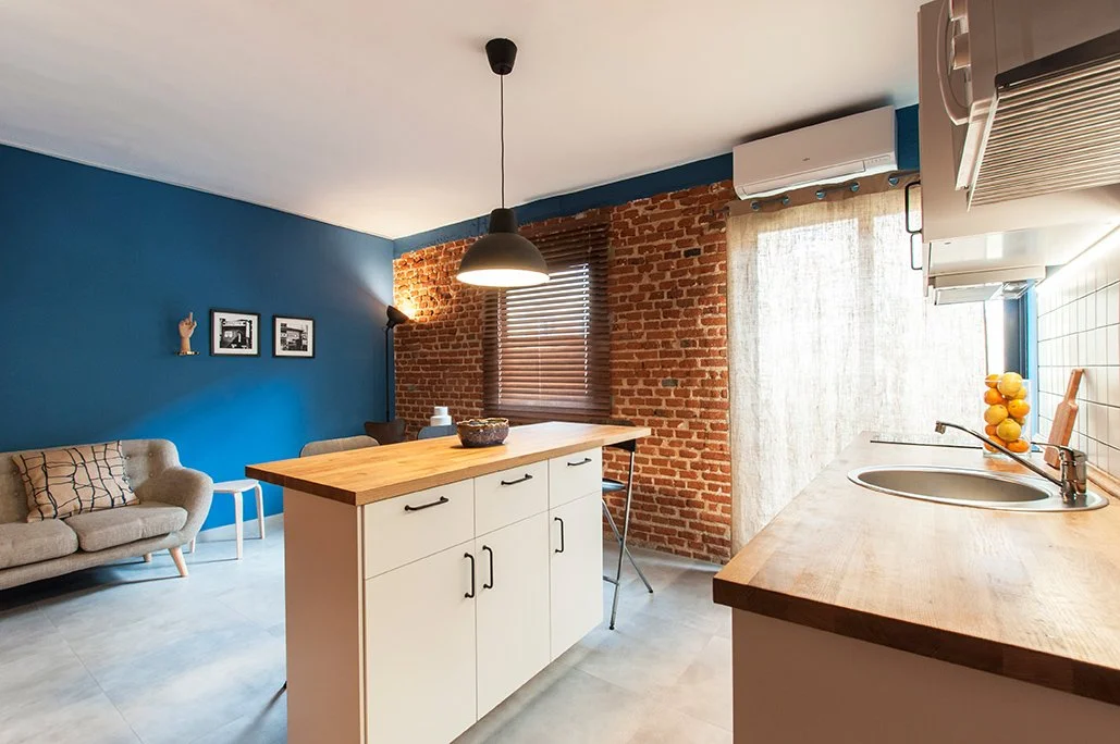 Interior de cocina moderna con paredes de ladrillo y azul, isla central de madera, sofá gris, y ventana con persianas de madera. Decoración minimalista.