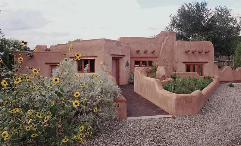 Casa estilo adobe con vegetación alrededor, incluyendo flores amarillas, en un entorno árido y cielo nublado.